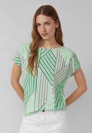 Frau mit langem hellbraunem Haar, die ein kurzärmeliges, grün-weiß gestreiftes Top mit geometrischem Muster und weiße Hosen vor einem neutralen Hintergrund trägt.