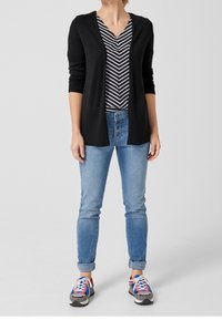 Lungo cardigan nero su una maglietta a righe bianche e nere con scollo a V, abbinato a jeans blu chiaro e sneakers multicolori.