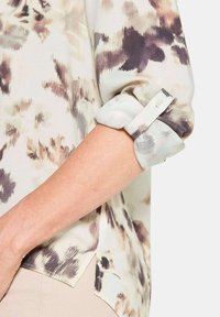 Blusa in tessuto leggero, con una base beige chiaro e motivi astratti marroni e grigi. Polsini con bottoni e maniche arrotolate.