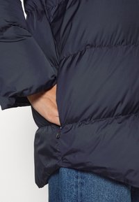 Marineblaue Steppjacke mit strukturiertem Obermaterial. Verfügt über eine seitliche Tasche mit Reißverschluss und elastischen Bündchen. Angezeigt mit Jeans.