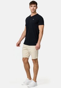 Camiseta negra de cuello en V, mangas cortas, pantalones cortos beige, zapatillas blancas. Diseño sencillo con un logo bordado en el pecho de la camiseta.