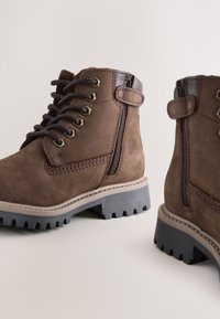 Braune Wildleder-Stiefeletten mit Schnür- und Reißverschluss, gummierten Profilsohlen und kontrastierenden Lederakzenten. Gestickte Details an den Seiten.