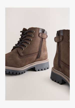 Pruunid seemisnahast pahkluu saapad paela- ja tõmblukuga kinnitusega, kummist kõrge haardega taldadega ning kontrastsed nahast detailid. Külgedel õmmeldud mustrid.