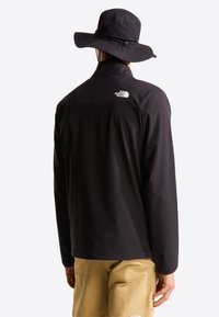 Μαύρο αδιάβροχο μπουφάν από ελαφρύ υλικό, με ψηλό γιακά και λογότυπο στην πλάτη. Συνδυάζεται με μπεζ σορτς.