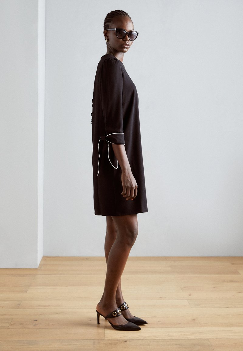 Black dress with three-quarter sleeves and white piping, paired with black pointed-toe heels featuring gold accents. Wood floor background.