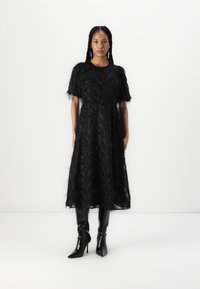 Robe noire avec un design texturé et en plumes, à manches courtes, associée à des bottes noires brillantes jusqu'aux genoux. Silhouette longue jusqu'au sol.
