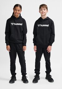 Zwei Kinder tragen passende schwarze Kapuzenpullis und Hosen, die ein weißes "hummel"-Logo aufweisen. Der Stoff wirkt weich und die Passform ist lässig.
