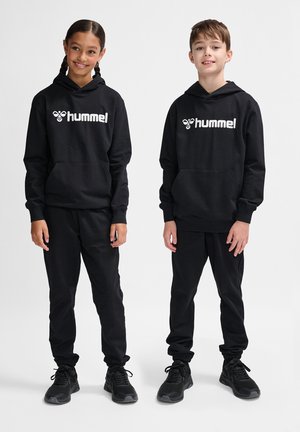 Twee kinderen dragen bijpassende zwarte hoodies en broeken, met een wit "hummel" logo. De stof lijkt zacht en de pasvorm is relaxed.