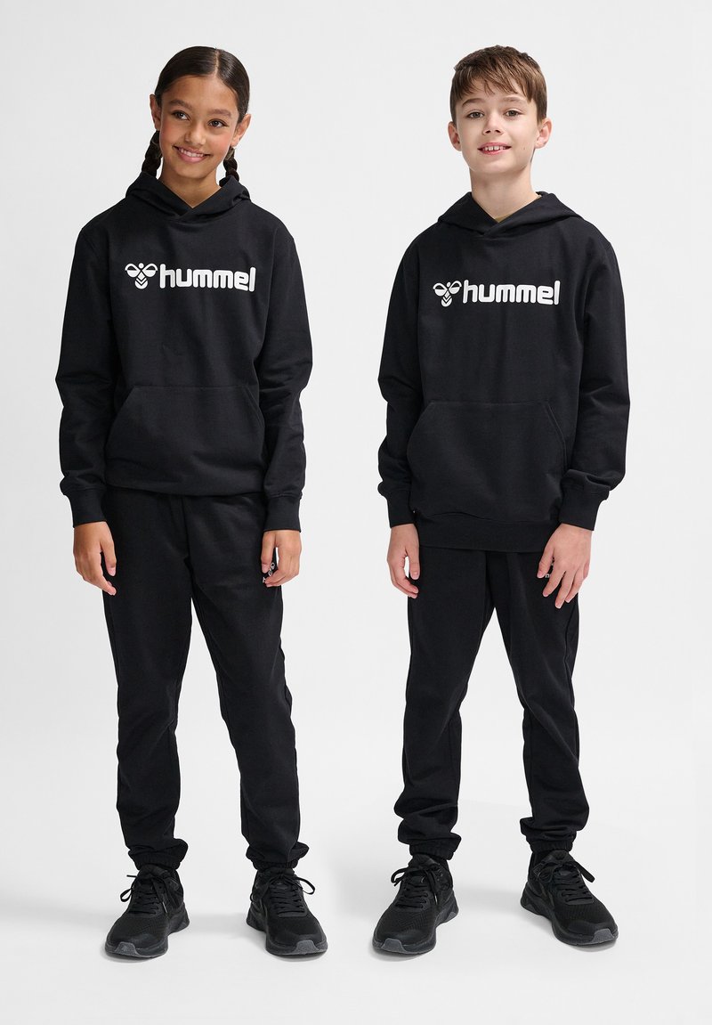 Zwei Kinder tragen passende schwarze Kapuzenpullis und Hosen, die ein weißes "hummel"-Logo aufweisen. Der Stoff wirkt weich und die Passform ist lässig.