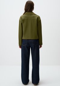 Veste vert olive avec un col, coupe boxy, et détail de couture au dos, associée à un jean en denim noir à jambe large.