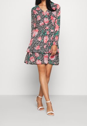 Blumenkleid mit langen Ärmeln, V-Ausschnitt und geschichteten Rüschen. Schwarzer Hintergrund mit pinken Rosen und grünen Blättern. Kombiniert mit weißen High Heels.