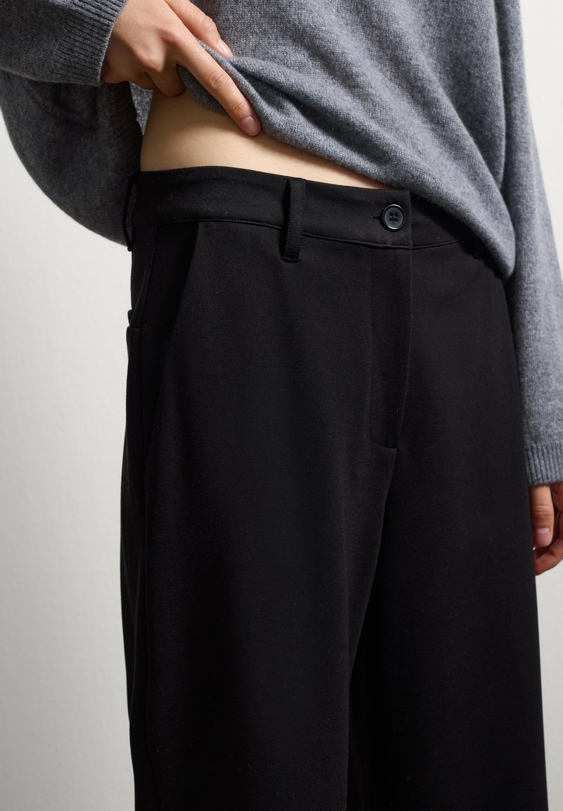 Zwarte broek met een gladde textuur, voorzien van een knoopsluiting, zijzakken en een getailleerd ontwerp. Gecombineerd met een grijze trui.