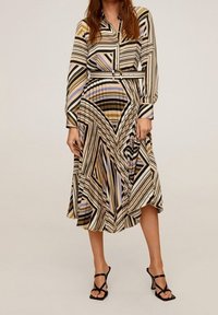 Femme portant une robe midi à manches longues, ceinturée, avec des rayures géométriques en beige, noir, blanc et bleu, associée à des sandales à talons noires à lanières.