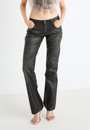 Pantalones de imitación cuero negros con corte recto, con bolsillos frontales y una textura sutil, combinados con sandalias de tacón transparente.