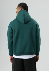 Donkergroene hooded trui met een relaxte pasvorm, gemaakt van zachte stof. Voorzien van geribbelde manchetten en zoom, gedragen over een wit shirt.