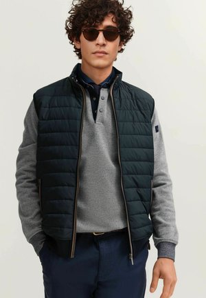 Man wearing dark sunglasses, black quilted vest over gray long-sleeve shirt, and navy trousers, standing against a plain light background.