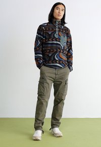 Fleece puloveris su abstrakčiu mėlynos, oranžinės ir rudos spalvų raštu, krūtinės kišenė žaliai; derinamas su alyvuogių spalvos krovinių kelnėmis ir šviesiai kreminiais sportbačiais.