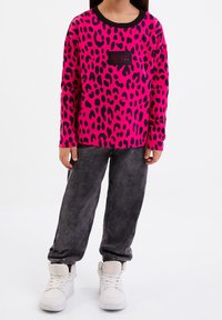 Sudadera de manga larga fucsia con estampado de leopardo negro, con un parche de logo negro, combinada con pantalones de estilo denim grises y zapatillas blancas.