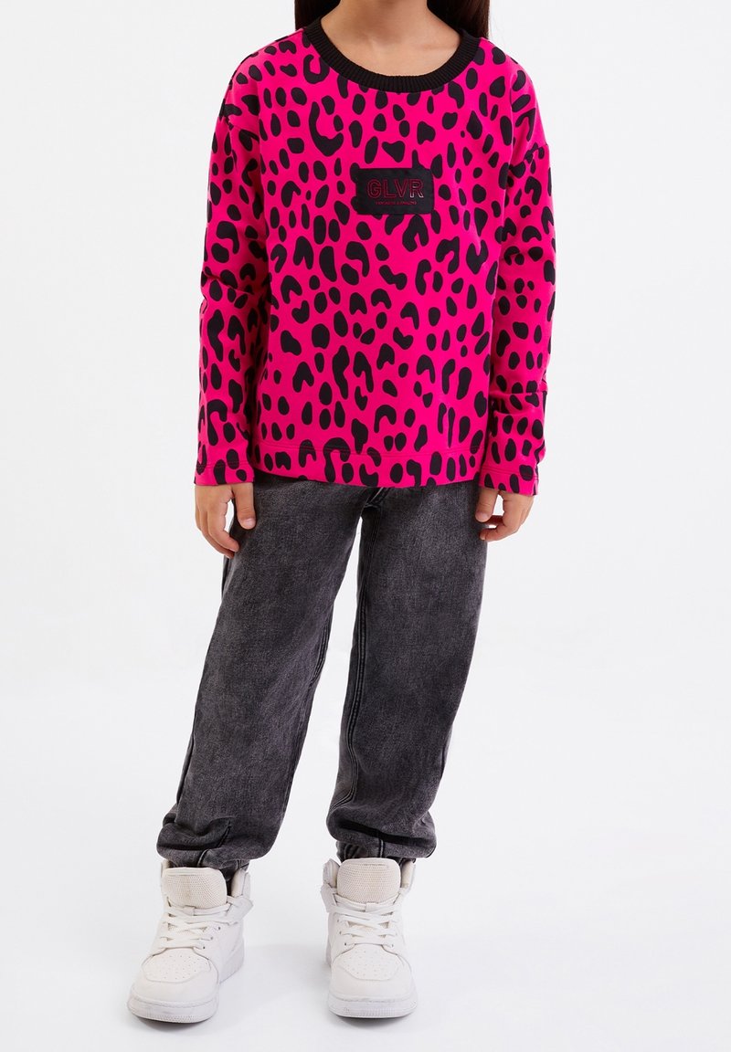 Sudadera de manga larga fucsia con estampado de leopardo negro, con un parche de logo negro, combinada con pantalones de estilo denim grises y zapatillas blancas.