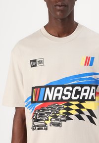T-shirt en coton beige avec un graphisme "NASCAR" en noir, accompagné de rayures colorées et d'illustrations de voitures de course, col rond, manches courtes.