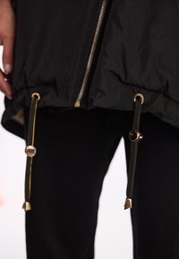 Czarna kurtka z złotym zamkiem i akcesoriami, z regulowanymi sznurkami na dole, wykonana z gładkiego, teksturowanego materiału, o luźnym kroju.