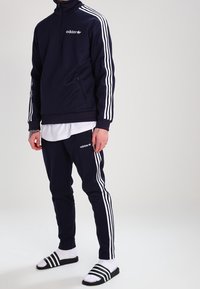 Chándal de color marino con diseño de tres rayas blancas en las mangas y los laterales, con cuello con cremallera y logo, combinado con sandalias tipo slide.