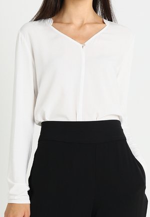 Blouse blanche à manches longues avec col en V rentrée dans un pantalon noir taille haute portée par une personne aux longs cheveux foncés, sur un fond uni.