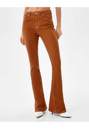 Slim-fit brown jeans with flared legs worn with white pointed-toe heels, model standing against a plain background.