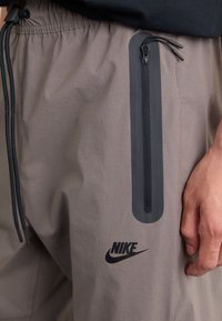 Pantaloni sportivi gri cu buzunar cu fermoar, talie elastică și logo-ul Nike de culoare neagră. Realizați dintr-un material ușor, neted, cu un finisaj mat.