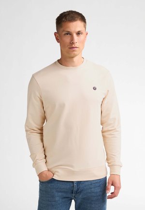 Jonge man met kort haar die een lichtbeige lange mouwen trui en blauwe spijkerbroek draagt, staand met één hand in de zak.