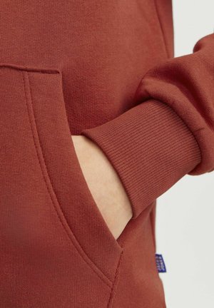 Zbliżenie na sweter w kolorze rdzy, z prążkowanym mankietem, teksturowanym materiałem i małą niebieską metką na bocznej szwie.