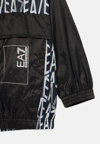 Μαύρο αδιαβροχο jacket από ελαφρύ υλικό, με ένα τολμηρό λευκό και ανοιχτό μπλε μοτίβο λογότυπου στο πλάι και ελαστικές μανσέτες.