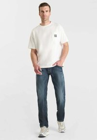 Uomo che indossa una camicia bianca a maniche corte con taschino, jeans blu e sneakers bianche, in piedi contro uno sfondo chiaro e semplice.