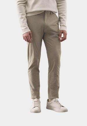 Mann trägt beige Slim-Fit-Hose, cremefarbene Sneaker und hellbeigen Langarm-Pullover, steht vor einem einfarbigen hellen Hintergrund.