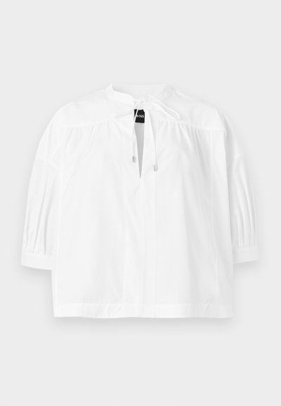 Blouse blanche à manches bouffantes trois-quarts, empiècement froncé et détail noué au col, présentée à plat sur un fond clair.