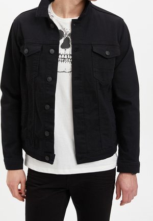 Mann trägt eine schwarze, geknöpfte Jeansjacke über einem weißen T-Shirt mit schwarzem Totenkopf-Print, kombiniert mit schwarzen Hosen, gezeigt von Hals bis Hüfte.