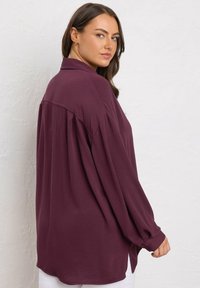 Donna con capelli castani che indossa una camicia lunga e larga di colore prugna, si volta per guardare oltre la spalla contro un muro bianco e testurizzato.