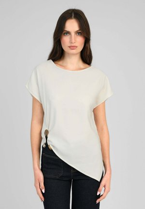 Femme aux longs cheveux foncés portant un haut asymétrique blanc à manches courtes avec un détail froncé sur le côté et un jean taille haute foncé sur fond gris.