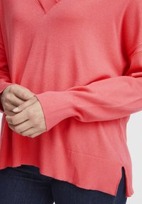 Maglione corallo a V con polsini e orlo a coste. Tessuto morbido, fessure laterali in basso e vestibilità comoda. Indossato con jeans scuri.