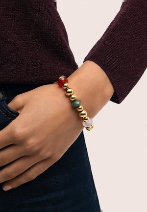 Pulsera de cuentas de oro y piedras preciosas que presenta cuentas rojas, verdes y blancas en un patrón alternado; llevada en una muñeca sobre una tela oscura.