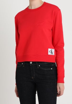 Czerwona krótka bluza z długim rękawem z naszywką z logo Calvin Klein w połączeniu z czarnymi jeansami z wysokim stanem na stojącej osobie.