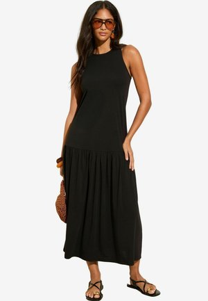 Abito maxi nero con design senza maniche, parte inferiore arricciata e tessuto liscio. Abbinato a sandali neri e accessori.
