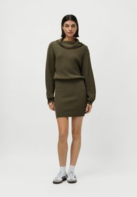 Frau, die in einem olivgrünen, gerippten Strickpullover mit Knopfdetails und passendem Minirock steht, trägt weiße Sneakers und Socken.