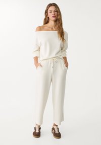 Bela off-the-shoulder sweatshirt v paru s ohlapnimi, usklajenimi hlačami. Ima elastičen pas, stranske žepke in rjave športne čevlje.