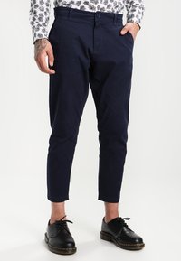 Mies pukeutuneena tummansinisiin slim fit -leikatuihin polvipituihin housuihin, valkoiseen paitaan, jossa on musta lehtikuvio, mustat nahkaiset nauhakenkä, seisomassa toinen käsi taskussa.