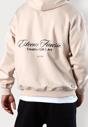 Person trägt hellbeigen Hoodie mit dem Text „Esteem Finesse Creative Craft & Art“ auf dem Rücken, kombiniert mit schwarzen Hosen und weißem Unterhemd.