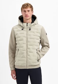 Man in een beige, gewatteerde jas met capuchon en textuursleeves, in zwarte broek, staand tegen een witte achtergrond met één hand in zijn zak.