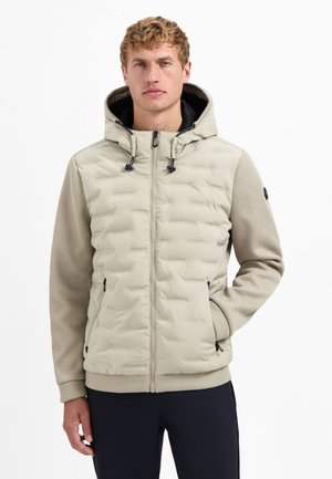 Man in een beige, gewatteerde jas met capuchon en textuursleeves, in zwarte broek, staand tegen een witte achtergrond met één hand in zijn zak.