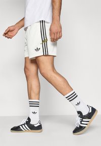 Pantalones cortos deportivos blancos con rayas negras, zapatos Adidas Samba negros y calcetas blancas hasta la rodilla con rayas negras y el logo.