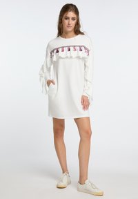 Weißes Kleid mit langen Ärmeln und Rüschendetails, einem dekorativen rosa Saum mit Quasten und einer lockeren Passform. Mit seitlichen Taschen.